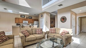 Ein geräumiges Wohnzimmer mit zwei beigefarbenen Sofas, einem Glastisch und einer Küche im Hintergrund.