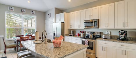 Una cocina con armarios blancos, encimera de granito, refrigerador de acero inoxidable, microondas, estufa y cafetera.