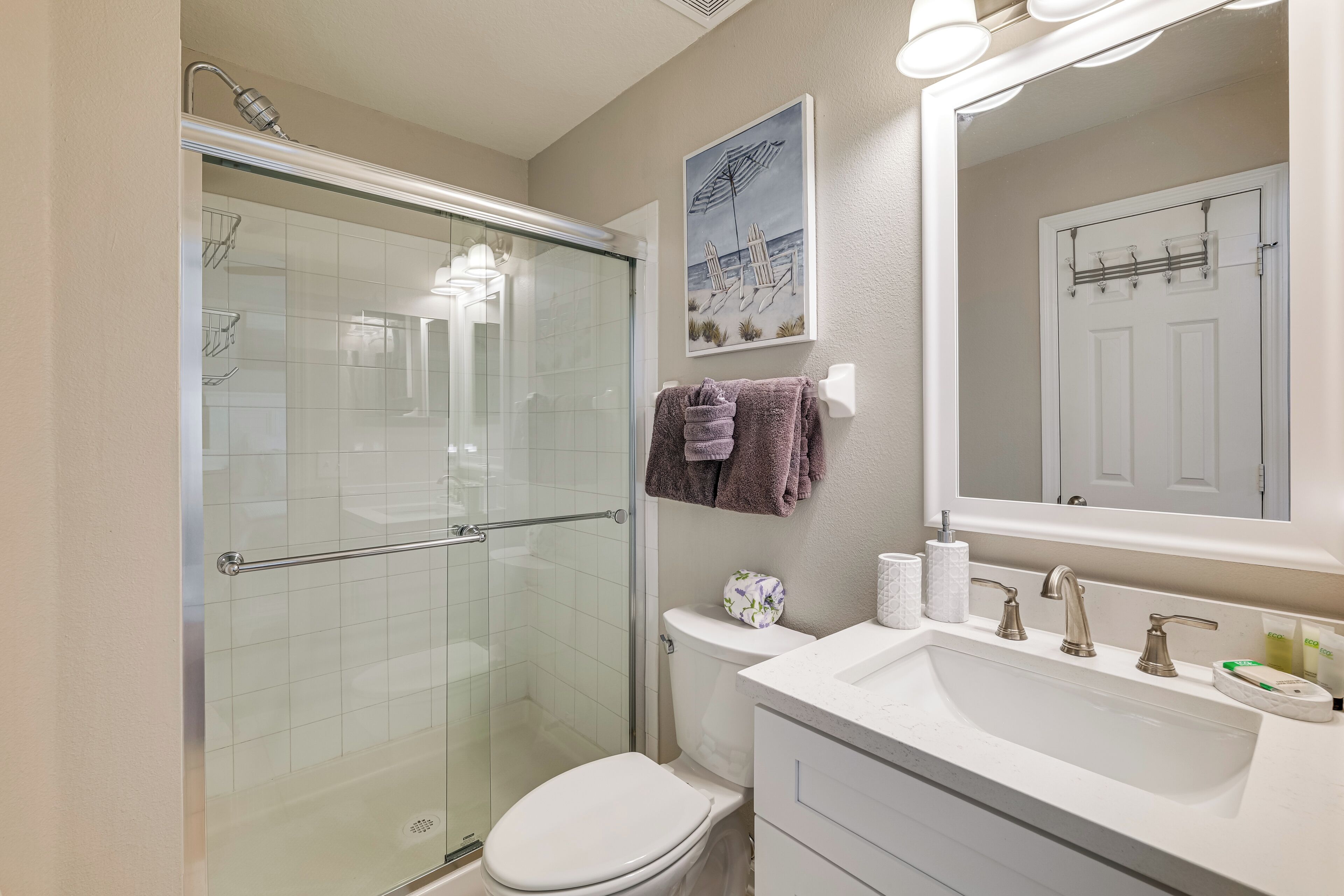 A bathroom with a glass shower enclosure, a white toilet, a sink with a mirror, and a towel rack with towels.