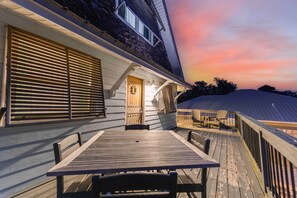 Eine Holzterrasse mit Tisch und Stühlen, ein überdachter Bereich mit Adirondack-Sesseln und ein Gebäude mit Natursteinfassade und Holzläden.