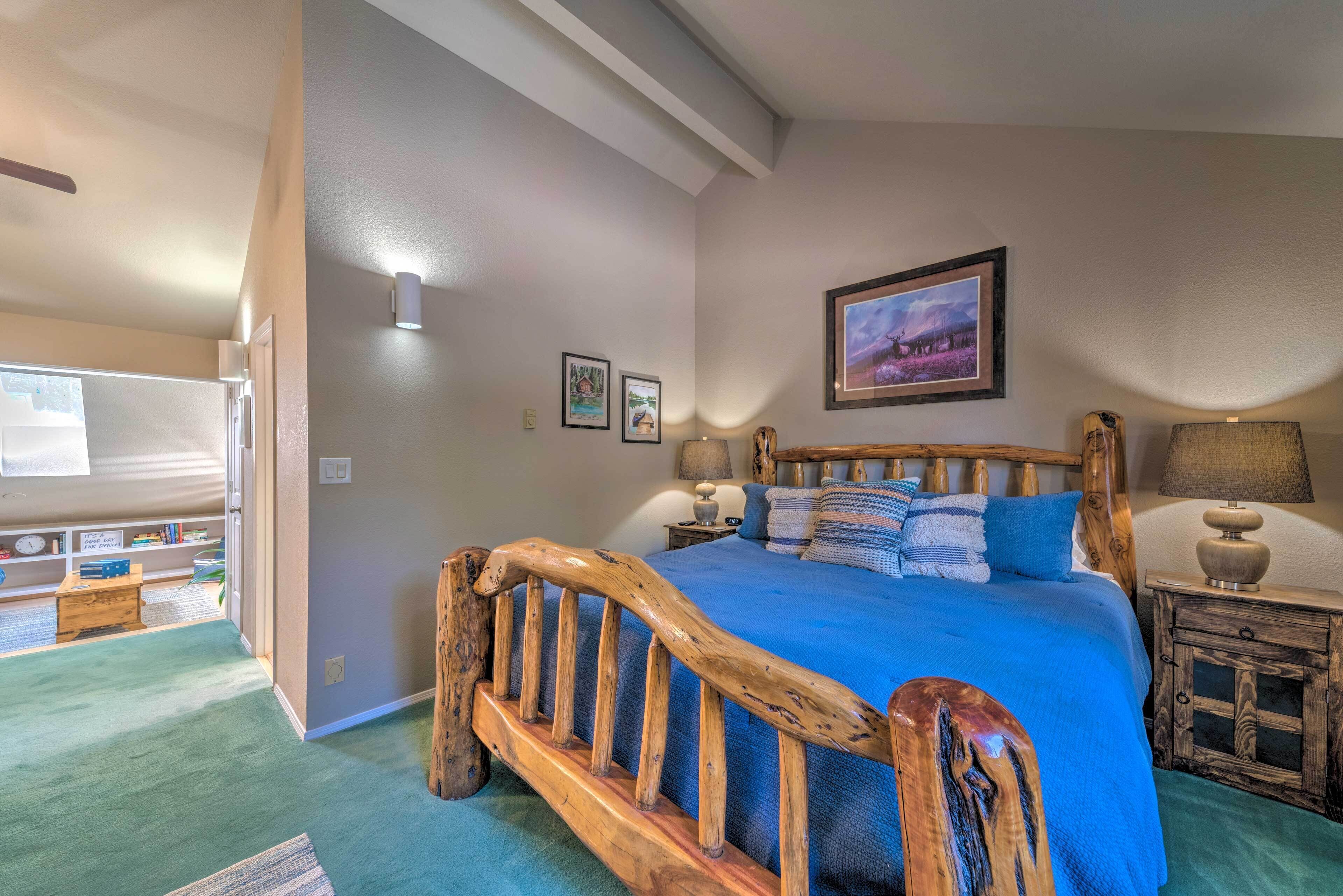 A bedroom with a wooden bed frame, a blue bedspread, and a nightstand with a lamp. There is a window with a view of the outdoors.
