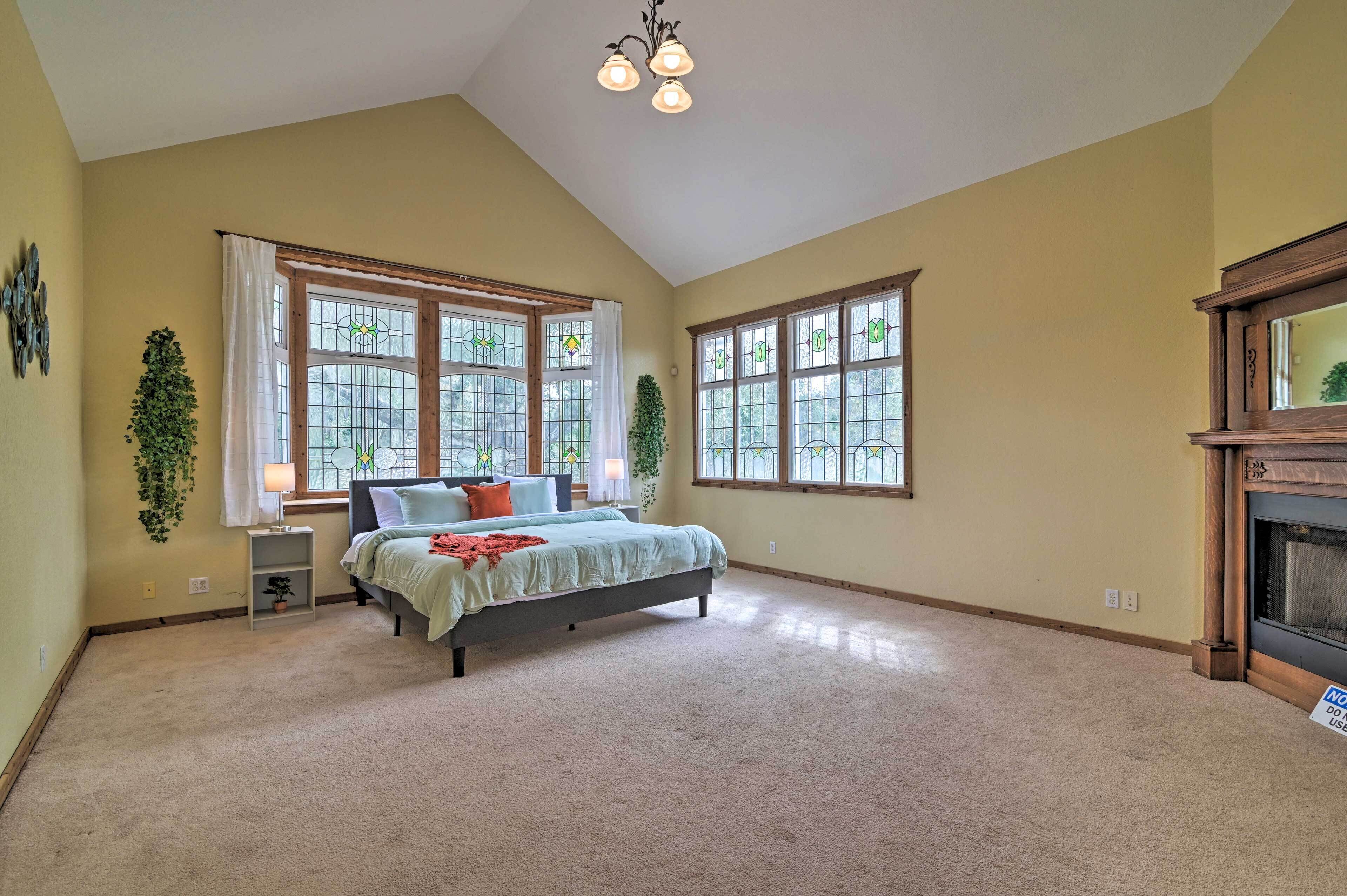 Un dormitorio espacioso con una cama grande, dos ventanas con cortinas, un candelabro y una chimenea.