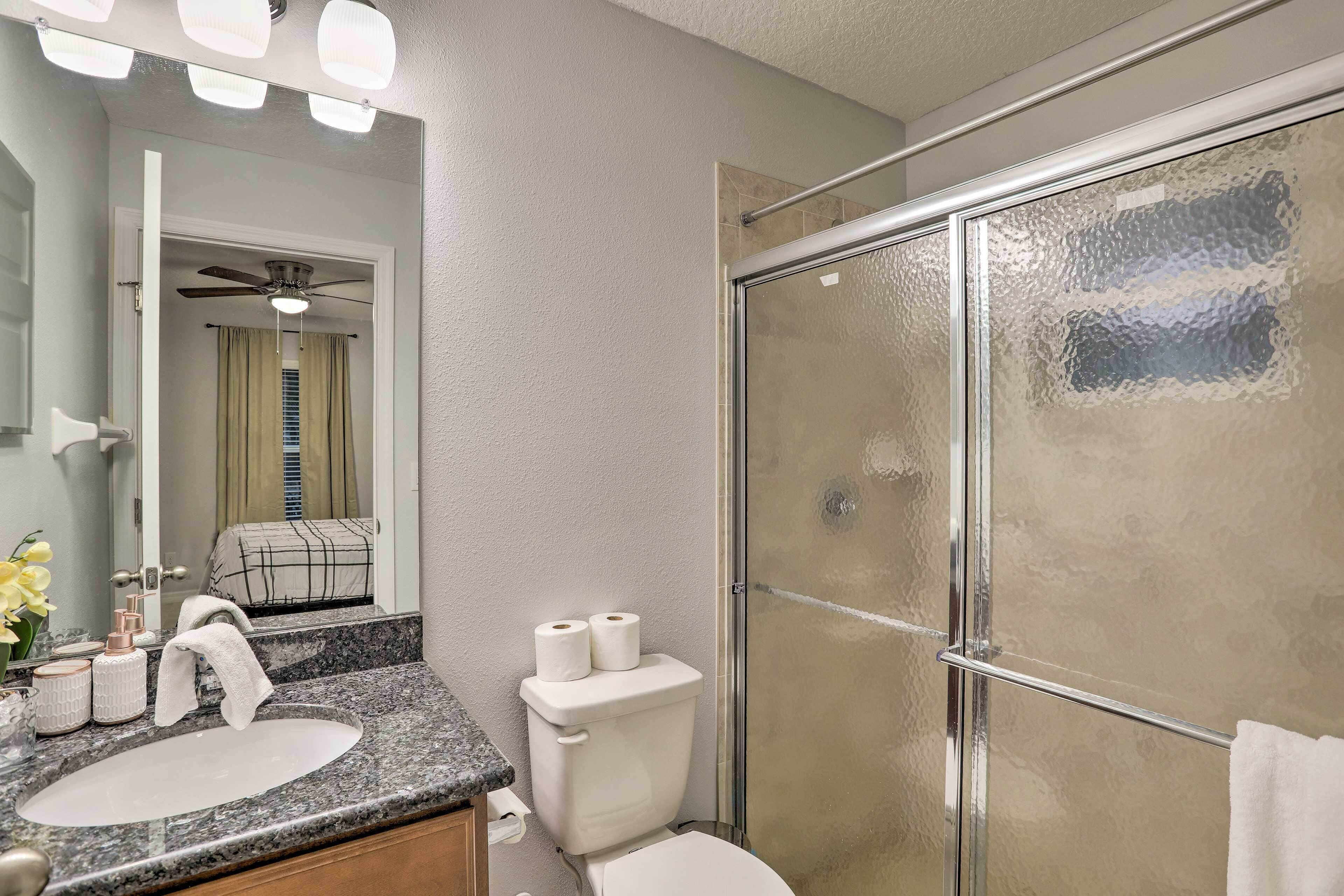 A bathroom with a granite countertop sink, a toilet with toilet paper, a glass shower enclosure, and a ceiling fan.
