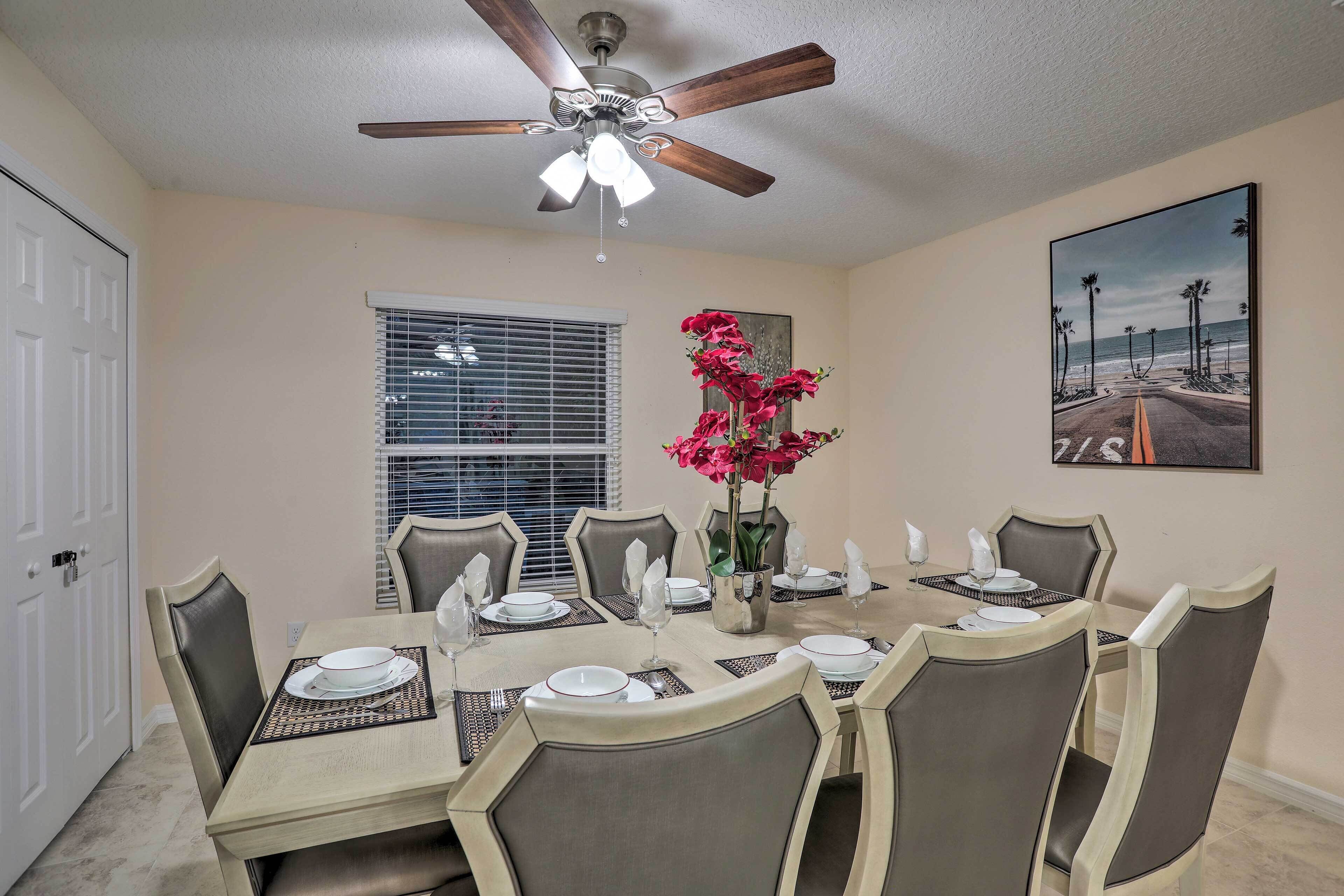 A dining area with a table set for six, a ceiling fan, a window with blinds, and a beach-themed painting on the wall.