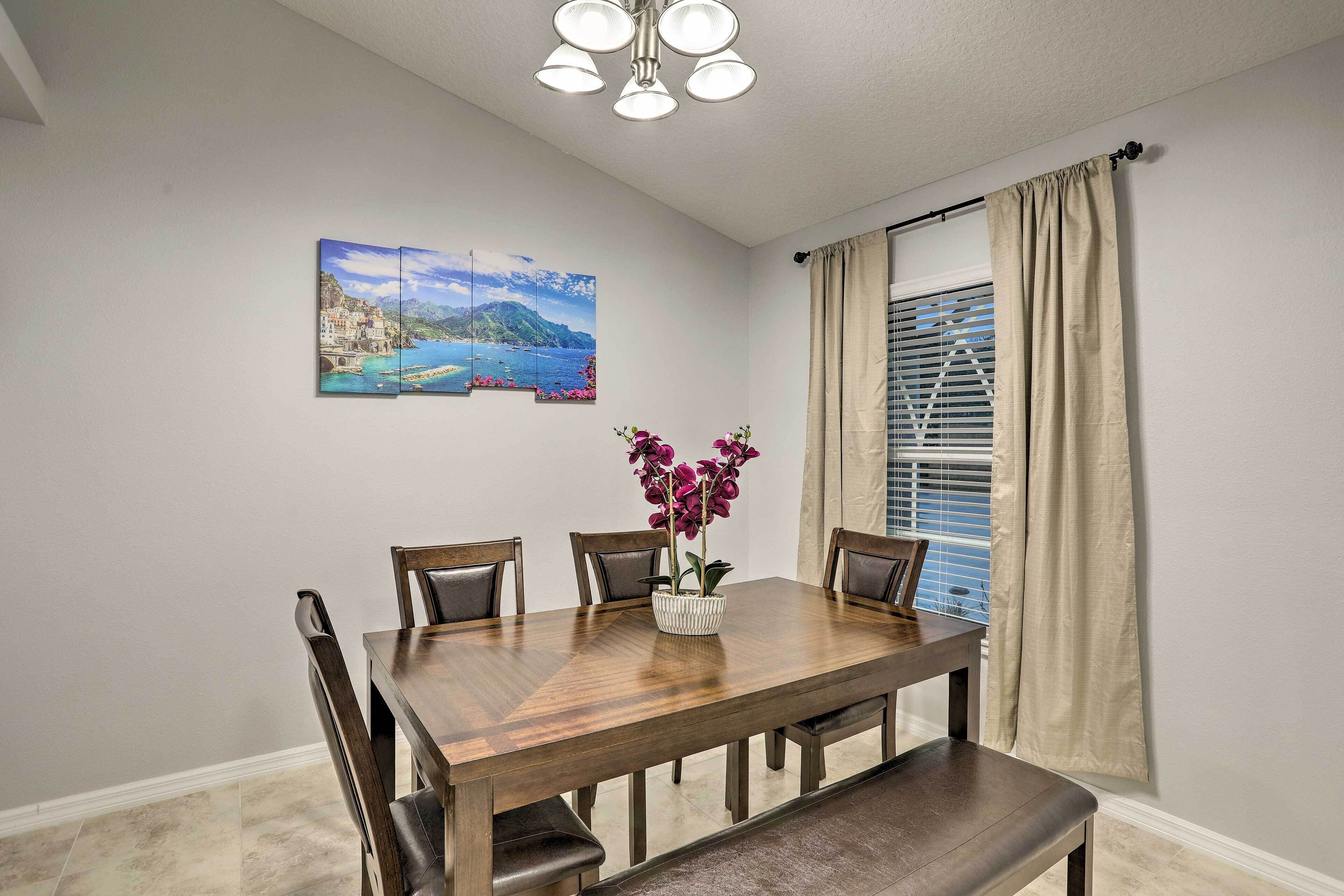 A dining room with a wooden table, four chairs, a vase with flowers, and a painting on the wall.