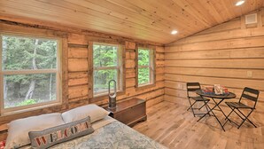 Une pièce de cabane en bois comprenant un lit, une petite table avec des chaises et de grandes fenêtres.