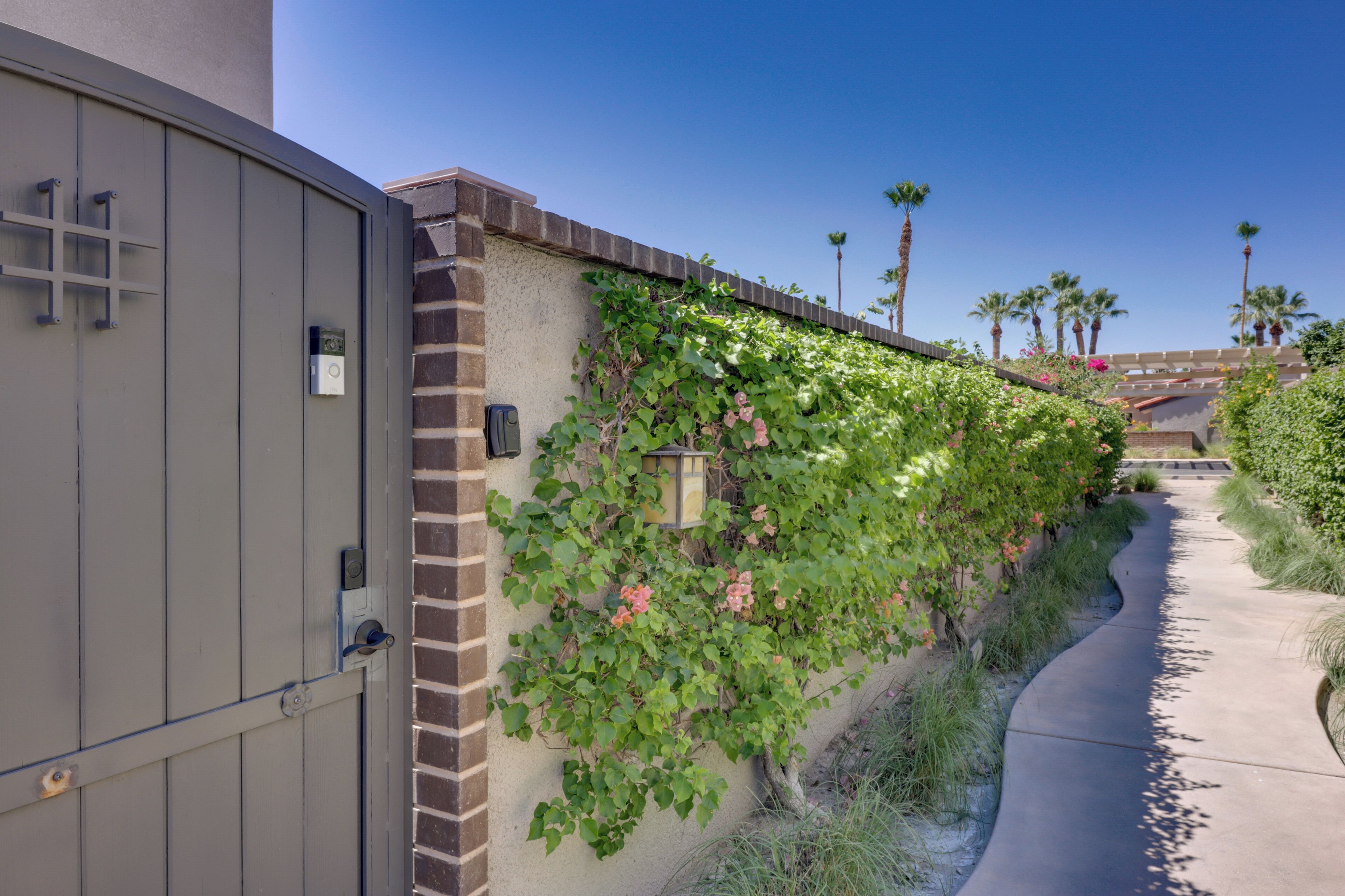 A gated entrance with a brick wall and climbing plants, a clear sky, and palm trees in the background.