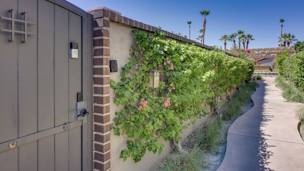 A gated entrance with a brick wall and climbing plants, a clear sky, and palm trees in the background. - Dtwn Palm Springs Condo: Bbq, Pool, Fire Pit, Etc! (Palm Springs)