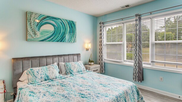 A bedroom with a bed, a headboard, a wall-mounted artwork, a bedside table with a lamp, and a window with curtains. - Fall Retreat at Barefoot Resort 1st Floor (North Myrtle Beach)