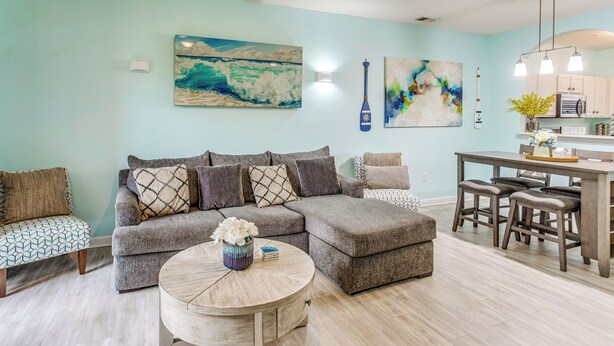 A living room with a sectional sofa, a round coffee table, and a dining area with a kitchen island. - Fall Retreat at Barefoot Resort 1st Floor (North Myrtle Beach)