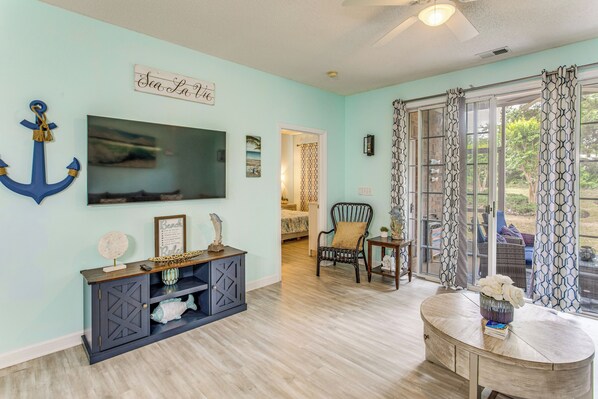 A room with a TV, a console table, a chair, and a view through a window. - Fall Retreat at Barefoot Resort 1st Floor (North Myrtle Beach)