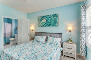 A bedroom with a bed, nightstands, a wall-mounted light, a door, and a bathroom visible in the background. - Luxury 1st-floor Barefoot Resort Condo! (North Myrtle Beach)
