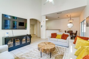 A modern living room with a flat-screen TV, a white sofa with colorful cushions, a wooden coffee table, and a dining area in the background.