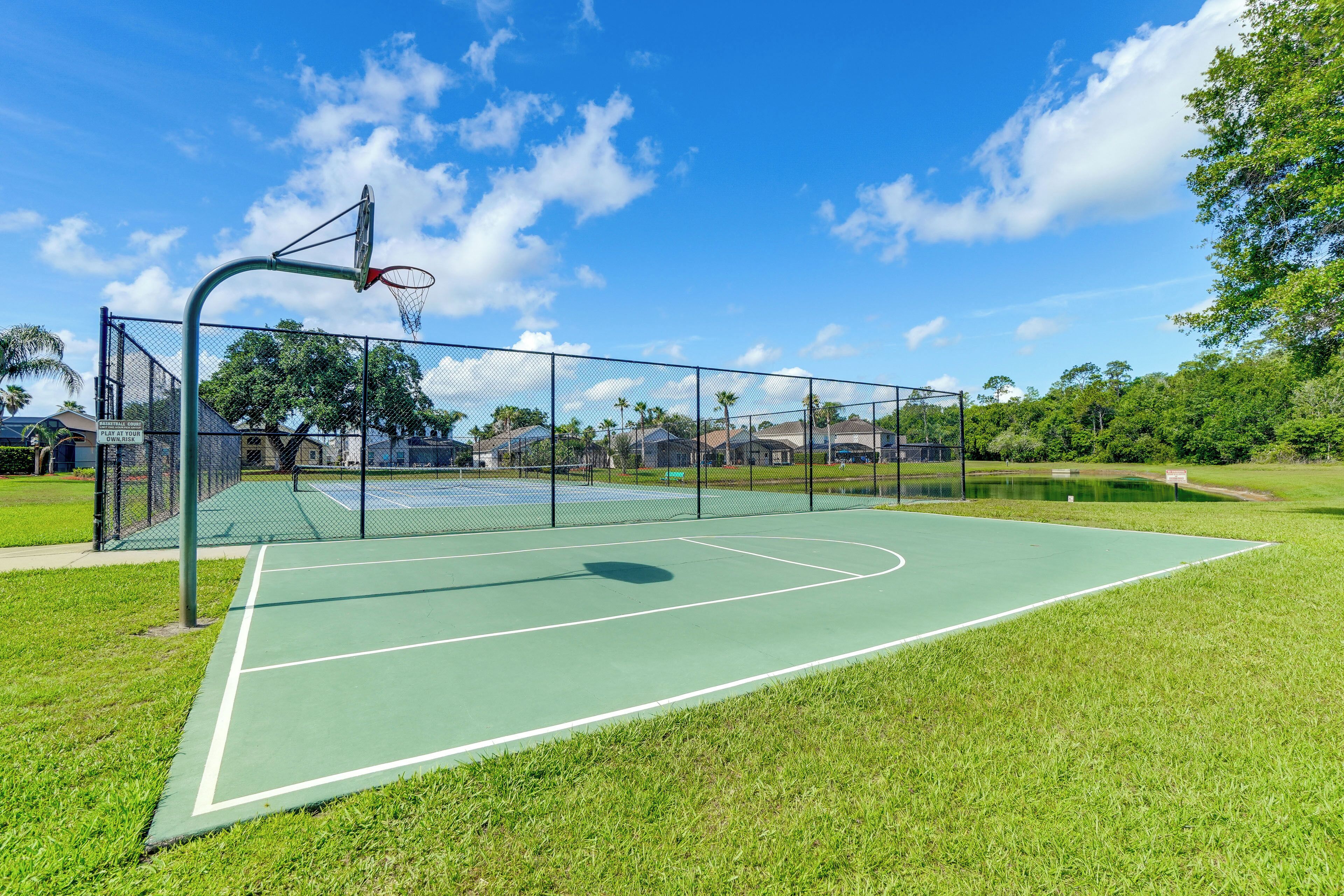An outdoor basketball court with a green surface, surrounded by a fence, and trees in the background.