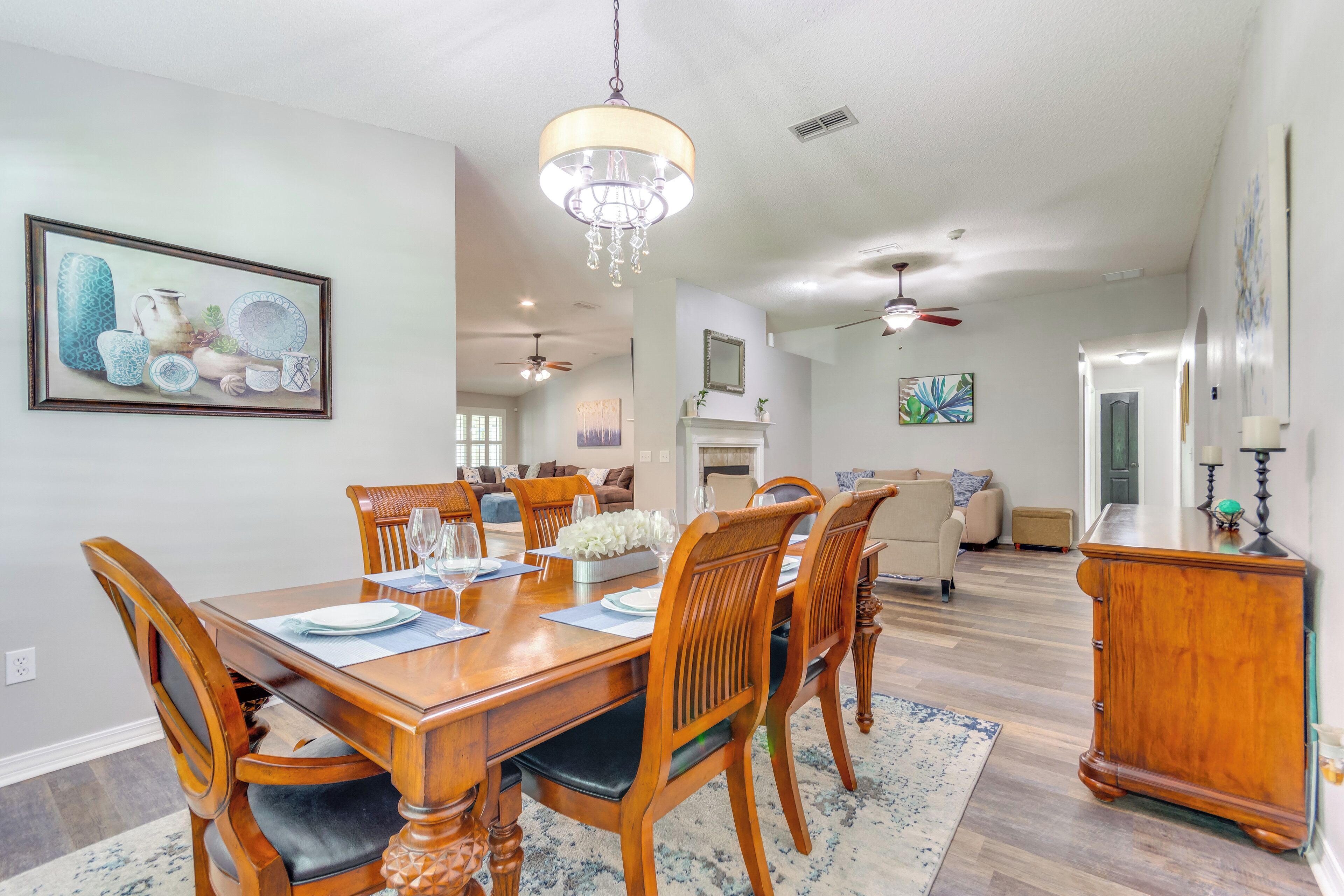 A dining area with a wooden table, chairs, and a chandelier. There is a painting on the wall and a fireplace in the background.
