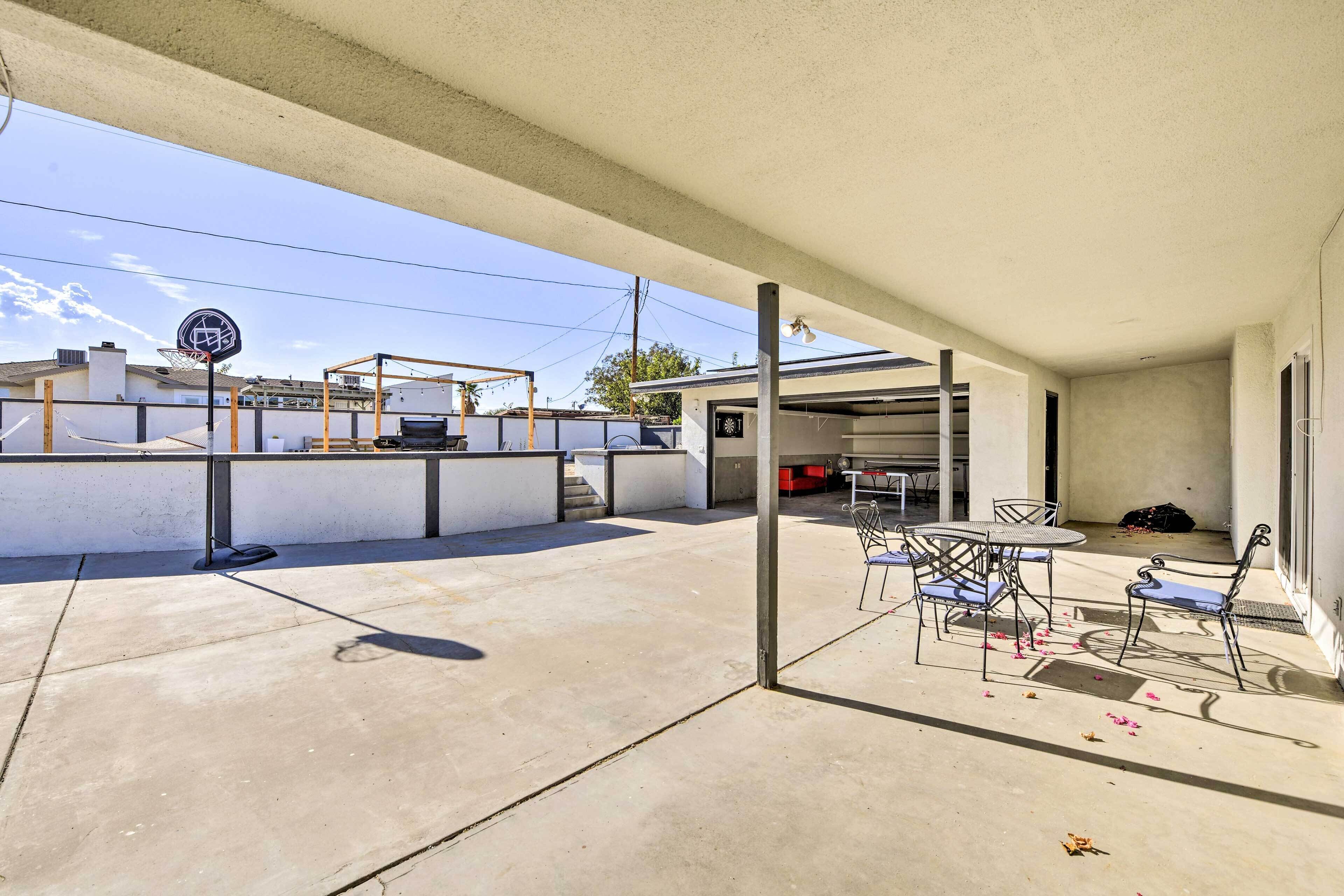 An outdoor patio area with a basketball hoop, a covered seating area with metal chairs and tables, and a view of a building in the background.