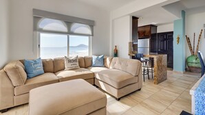 A spacious living room with a large window, a beige sectional sofa, a kitchen area, and a ceiling fan. - Beachfront Oasis in Cabo w/ Resort Amenities! (San José del Cabo)