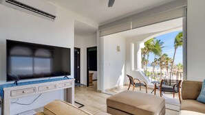 A modern living room with a flat-screen TV, beige sofas, and a balcony with a view of palm trees and the beach. - Beachfront Oasis in Cabo w/ Resort Amenities! (San José del Cabo)