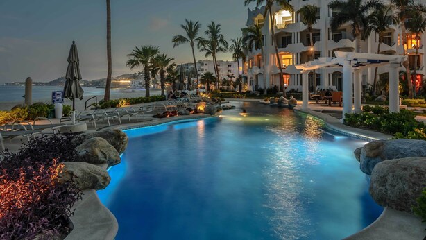 A resort pool with a view of the ocean, surrounded by palm trees and a white building with balconies. - Beachfront Oasis in Cabo w/ Resort Amenities! (San José del Cabo)