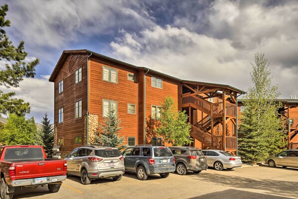 Un edificio de varios pisos con fachada de madera, un área exterior cubierta con escaleras y un estacionamiento con varios vehículos.