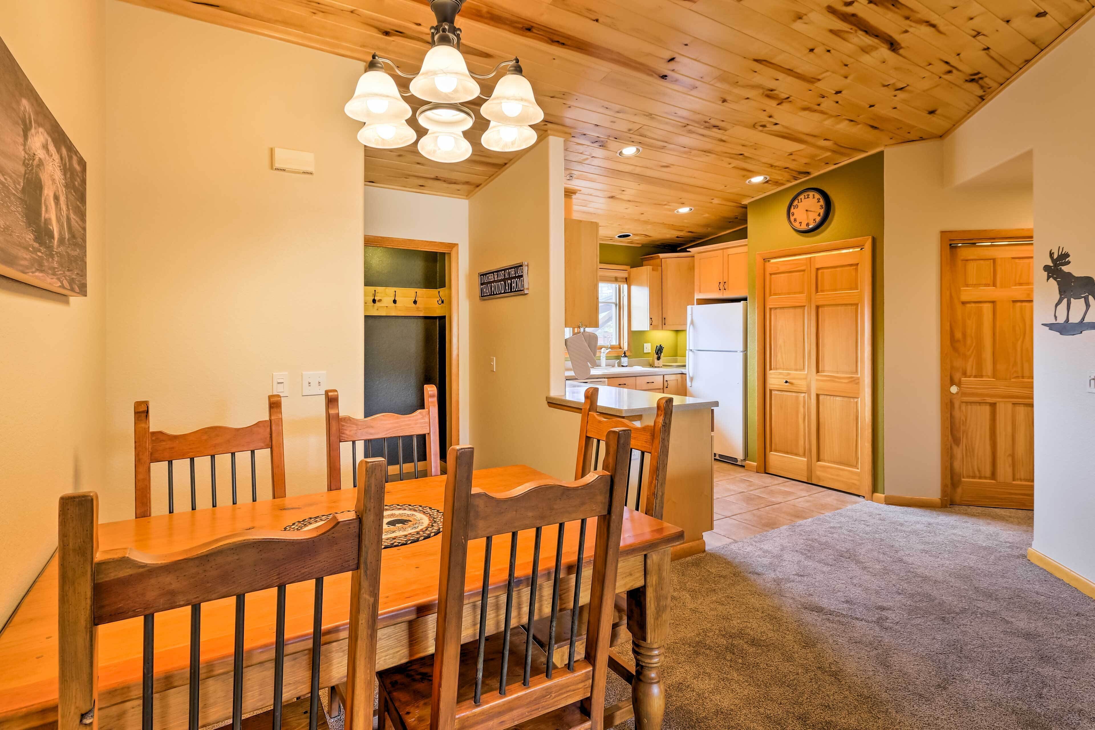 A dining area with a wooden table and chairs, a chandelier, and a kitchen in the background.