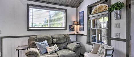 A cozy living room with a grey sofa, a small side table, a chair, and a window with blinds.