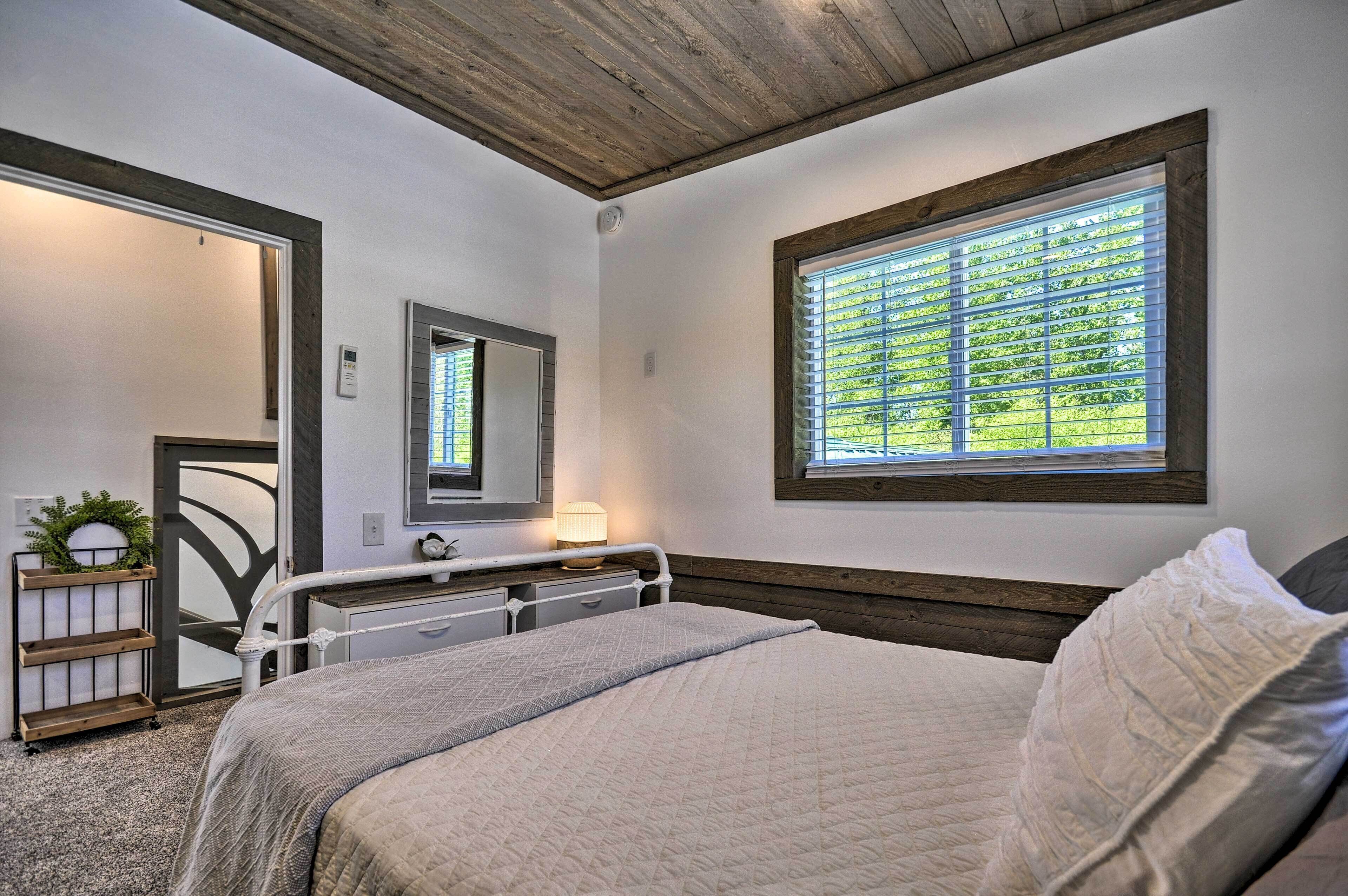 A bedroom with a bed, a bedside table with a lamp, a window with blinds, and a mirror on the wall.