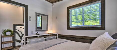 A bedroom with a bed, a bedside table with a lamp, a window with blinds, and a mirror on the wall.