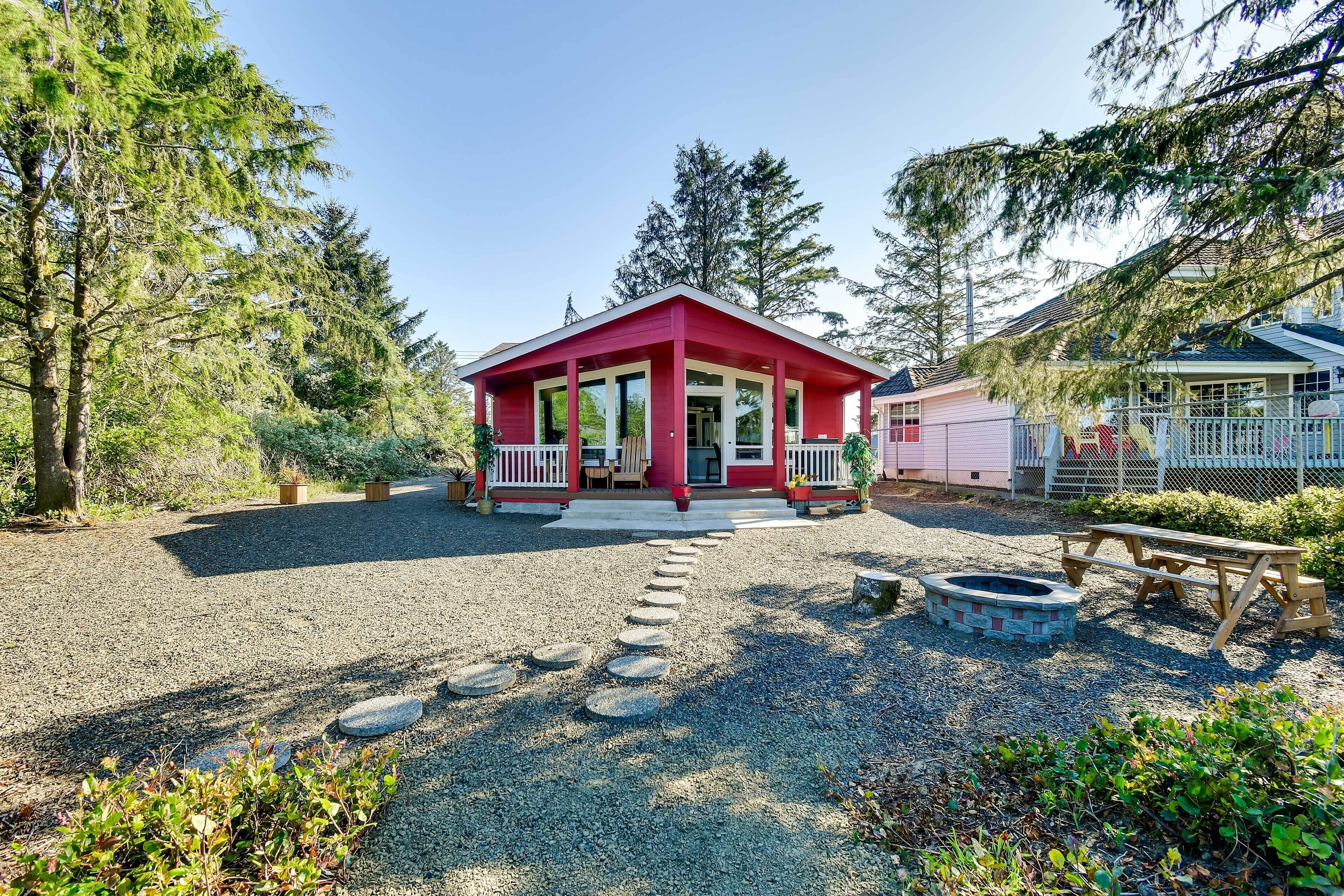 A red cabin with a porch, surrounded by trees and a gravel path leading to a fire pit.