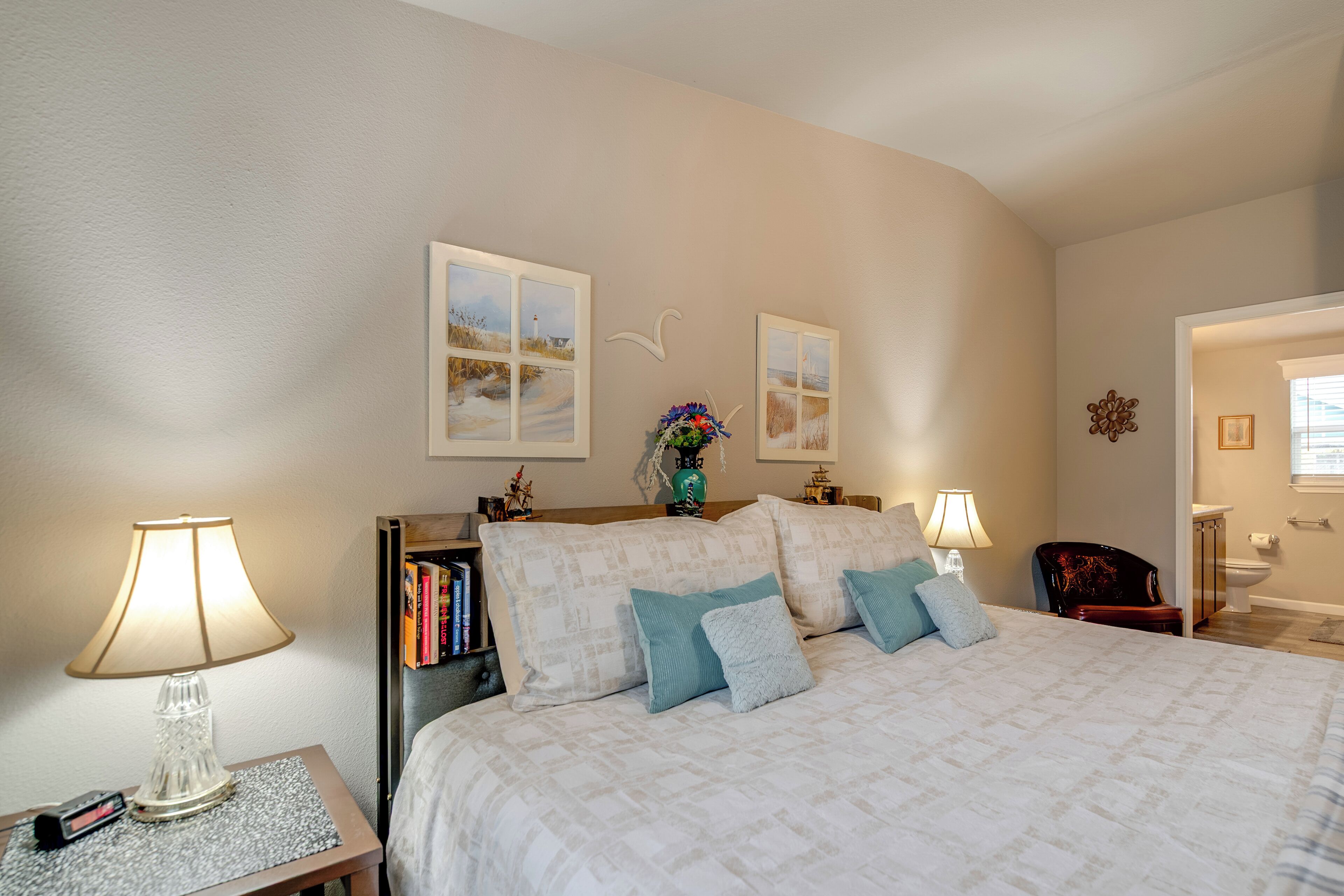 A bedroom with a large bed, two bedside lamps, a nightstand, a bookshelf, and a decorative wall piece.