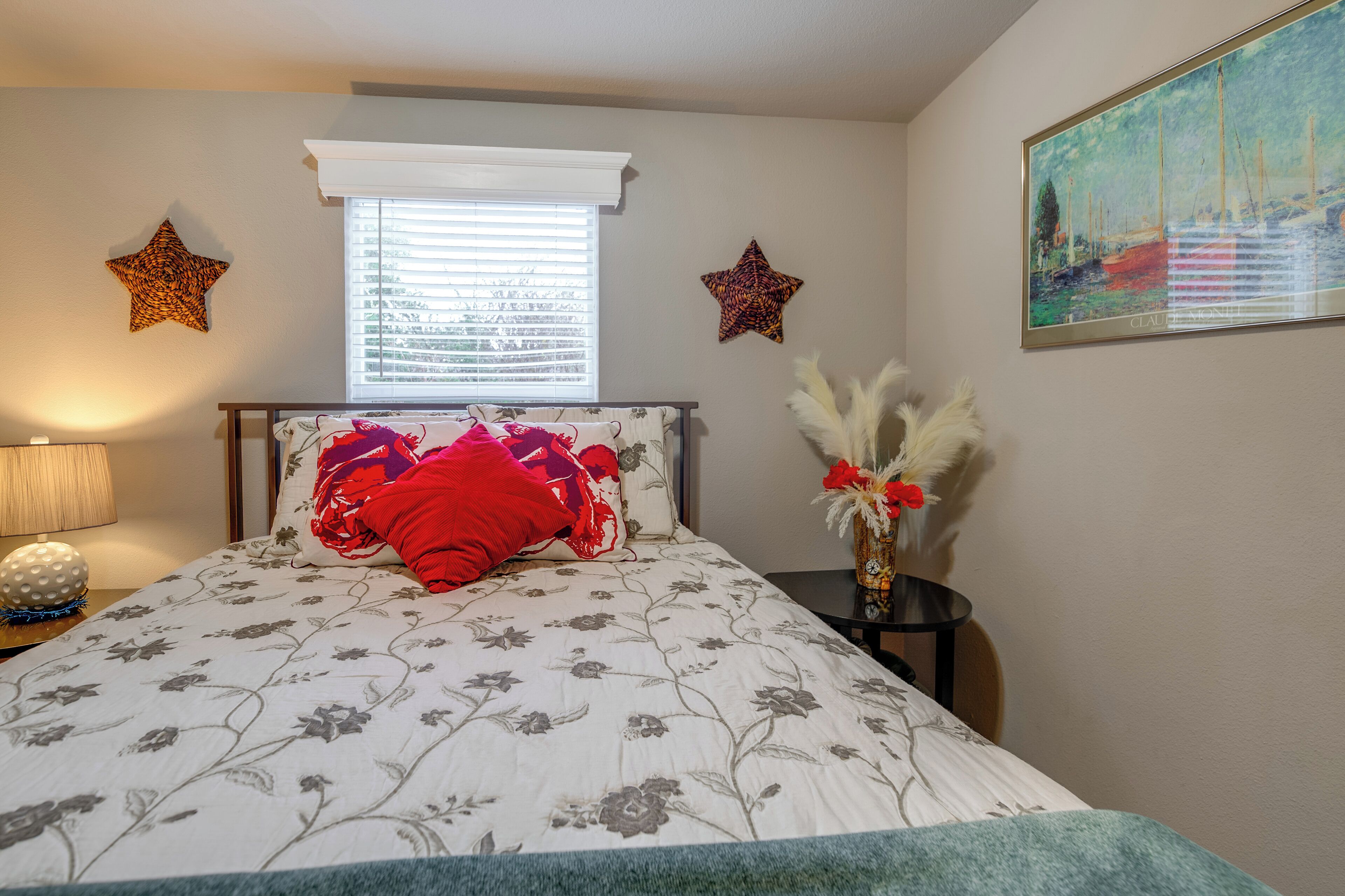 A bedroom with a bed, a nightstand, a lamp, a window with blinds, and two wall decorations.