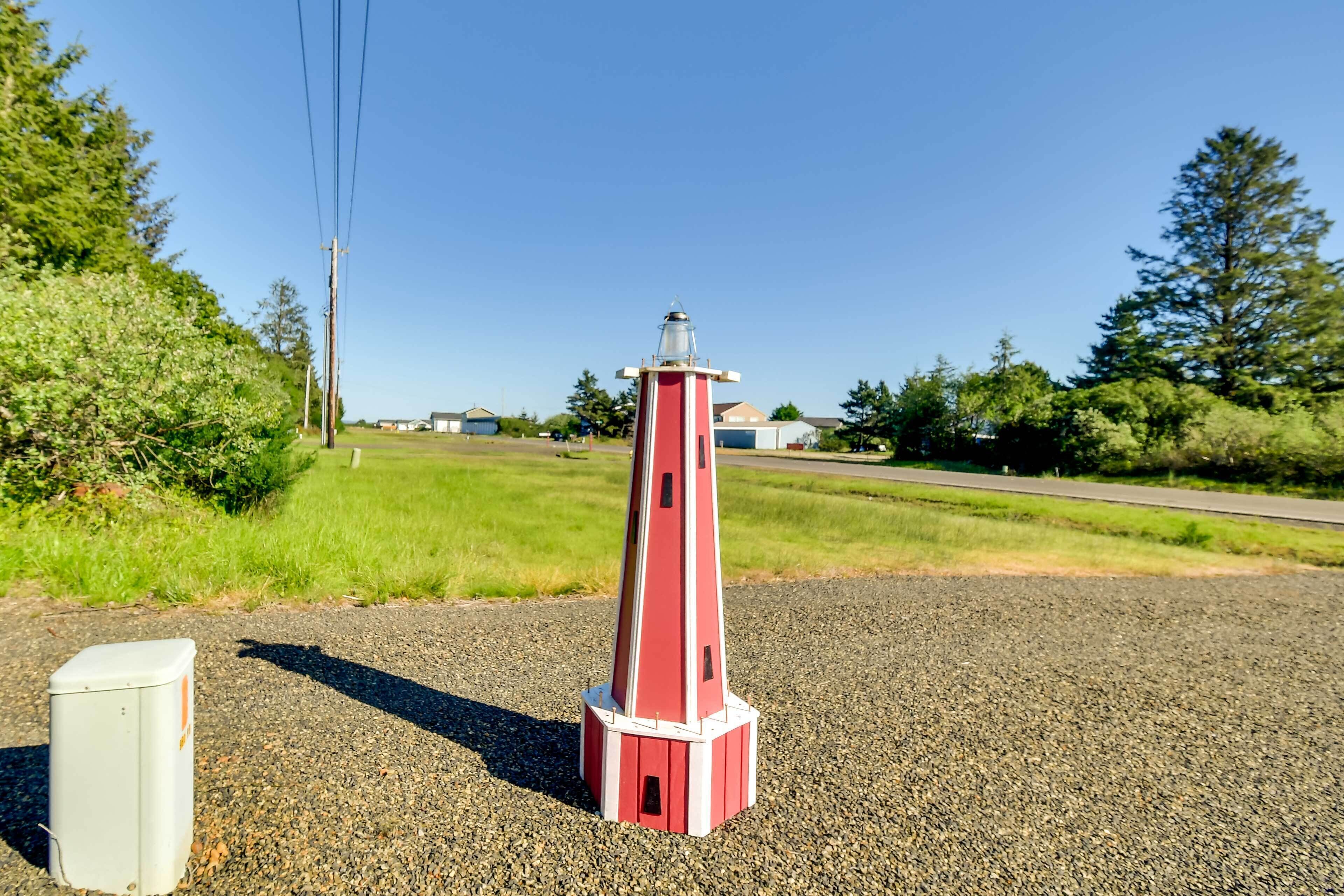 Un piccolo modello di faro rosso e bianco su uno sfondo di ghiaia, con un cielo sereno e vegetazione sullo sfondo.