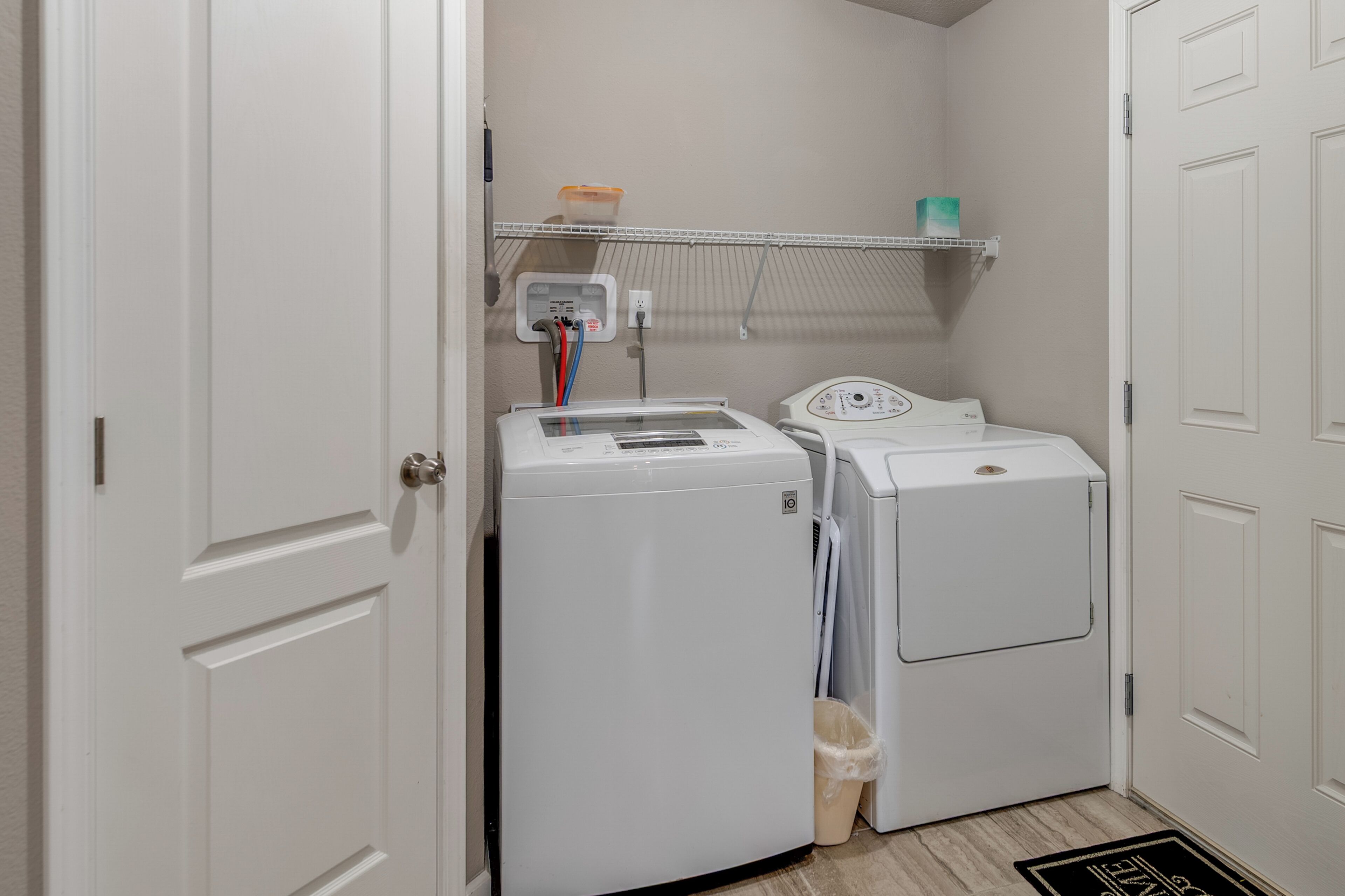 A laundry room with two white washing machines, a shelf with laundry supplies, and a door.