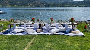 Mobiliario exterior para comer, con mesas y sillas, una manta y almohadas sobre un césped junto a un lago.