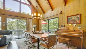 A spacious room with a wooden dining table, chairs, a piano, and a large window offering a view of trees. - Lake Arrowhead Family Home w/ Game Room (Lake Arrowhead)