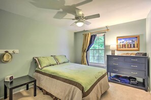Quarto com cama, penteadeira, janela com cortinas, ventoinha no teto e porta-chapéus fixado na parede.