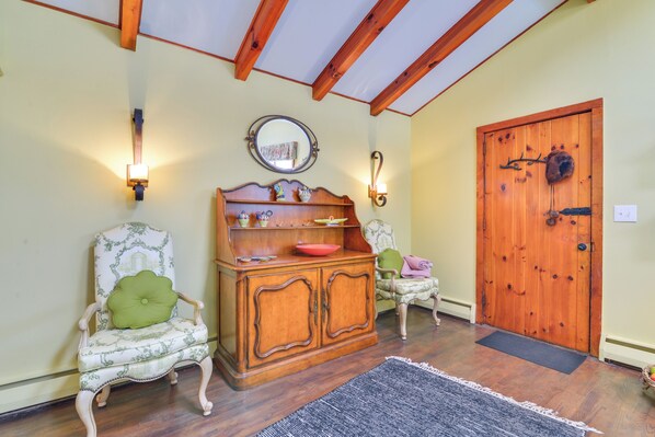 Une pièce avec un plancher en bois, une porte en bois, deux fauteuils rembourrés avec des coussins verts, un meuble en bois surmonté d’un miroir et un tapis noir sur le sol.