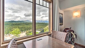 A room with a large window offering a mountain view, a wooden table, a woven chair, and a wall-mounted lamp.