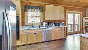 A wooden cabin kitchen with a stainless steel refrigerator, a dishwasher, wooden cabinets, a granite countertop, and a window with a curtain.
