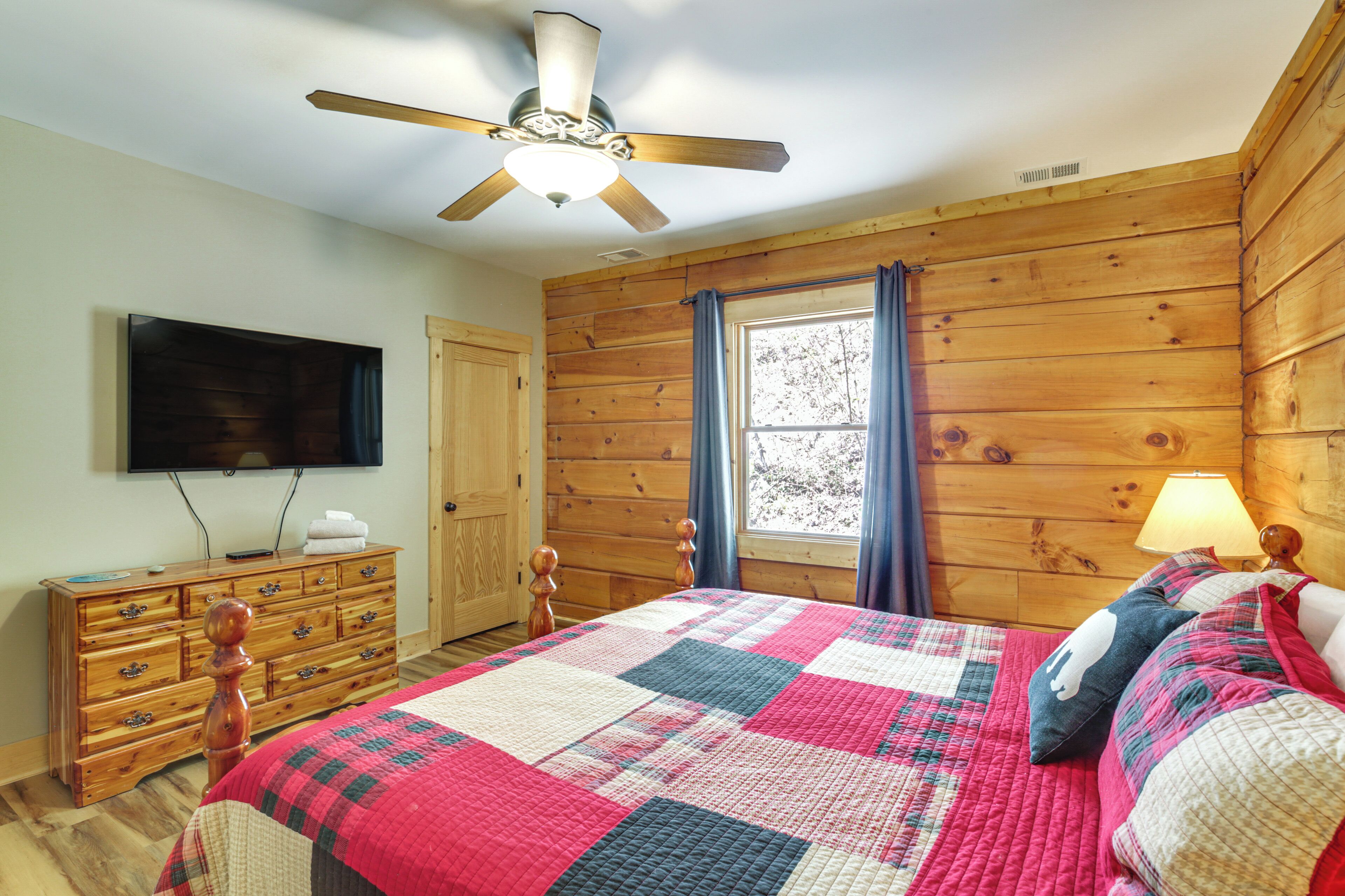 Une chambre avec un ventilateur de plafond en bois, une grande fenêtre avec des rideaux, une commode en bois et une couvre-lit matelassé.