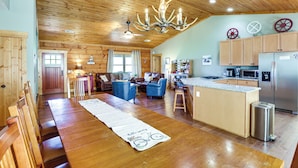 A spacious living area with a wooden ceiling, a chandelier, a dining table with chairs, and a kitchen area with a refrigerator and microwave.