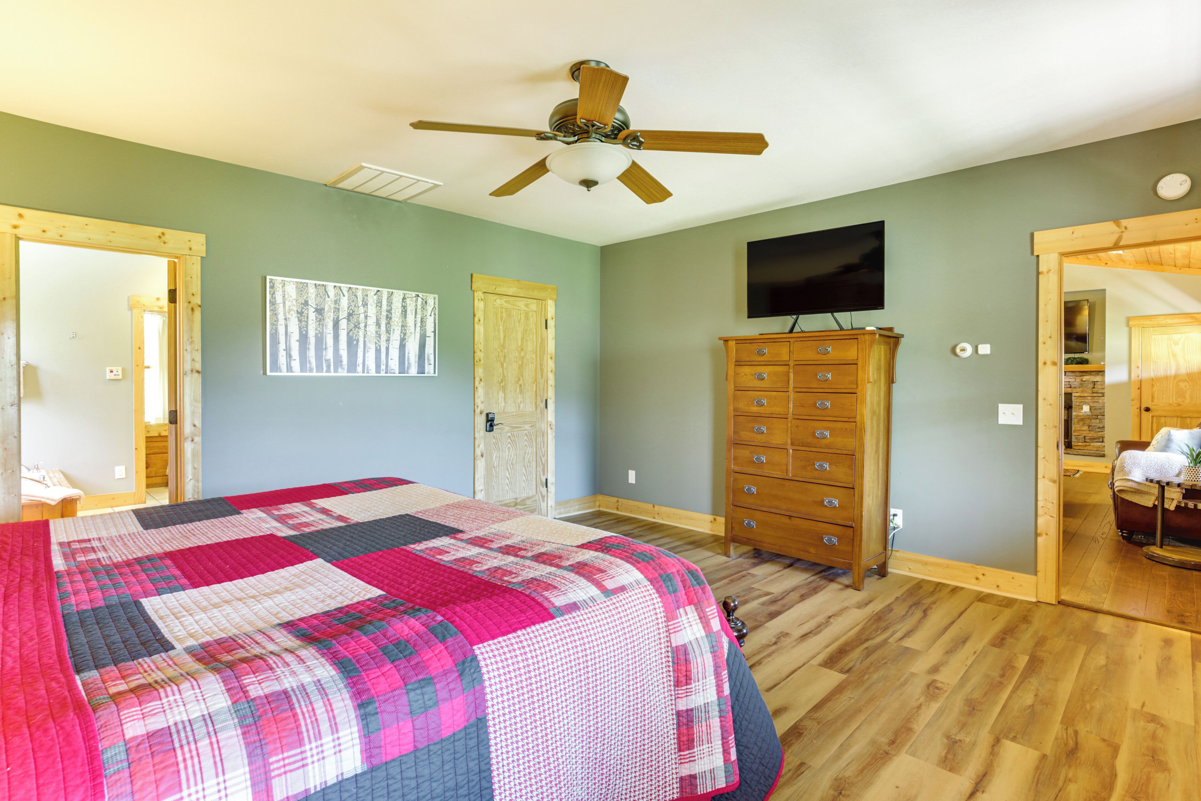 A bedroom with a large bed, a wooden dresser, a ceiling fan, a television, and a door leading to another room.