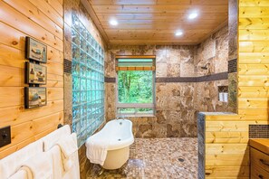 Un baño de cabaña de madera con bañera, ventana con vistas a follaje, y zona de ducha con azulejos.