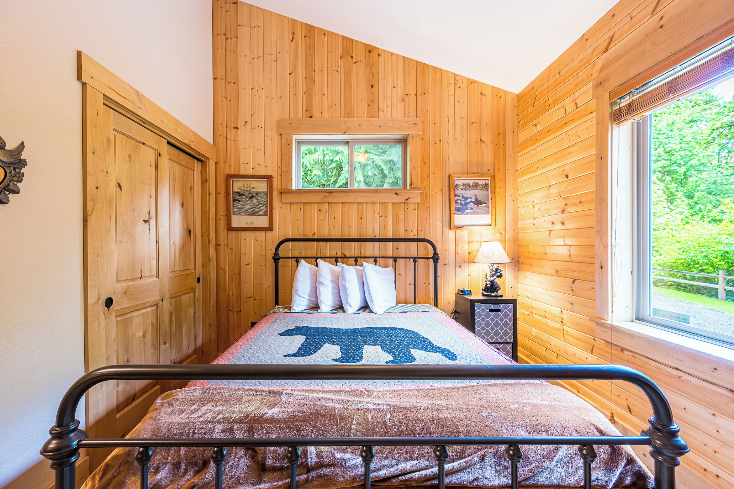 A wooden-paneled room with a bed, a window, and a small table with a lamp.