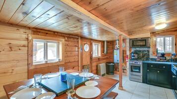 Interior de una cabaña de madera con comedor, cocina y un ventanal grande.