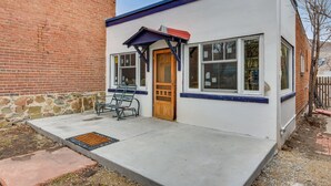 A small building with a brick wall, a wooden door, and a concrete porch with two chairs. - 0601: Downtown Salida Cottage w/ Fireplace + Yard (Salida)