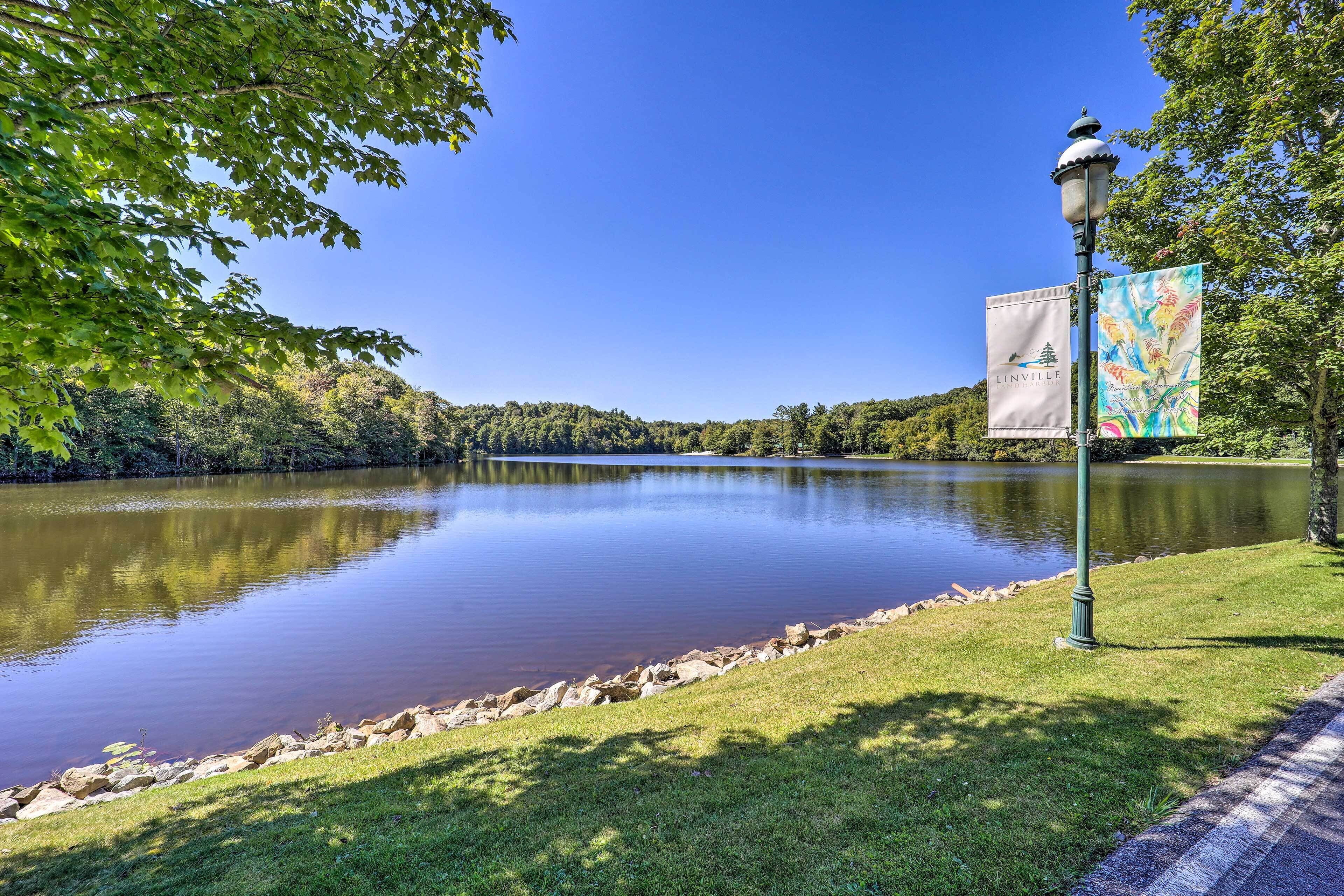 A calm lake with a clear sky, surrounded by lush greenery and a lamp post with banners.