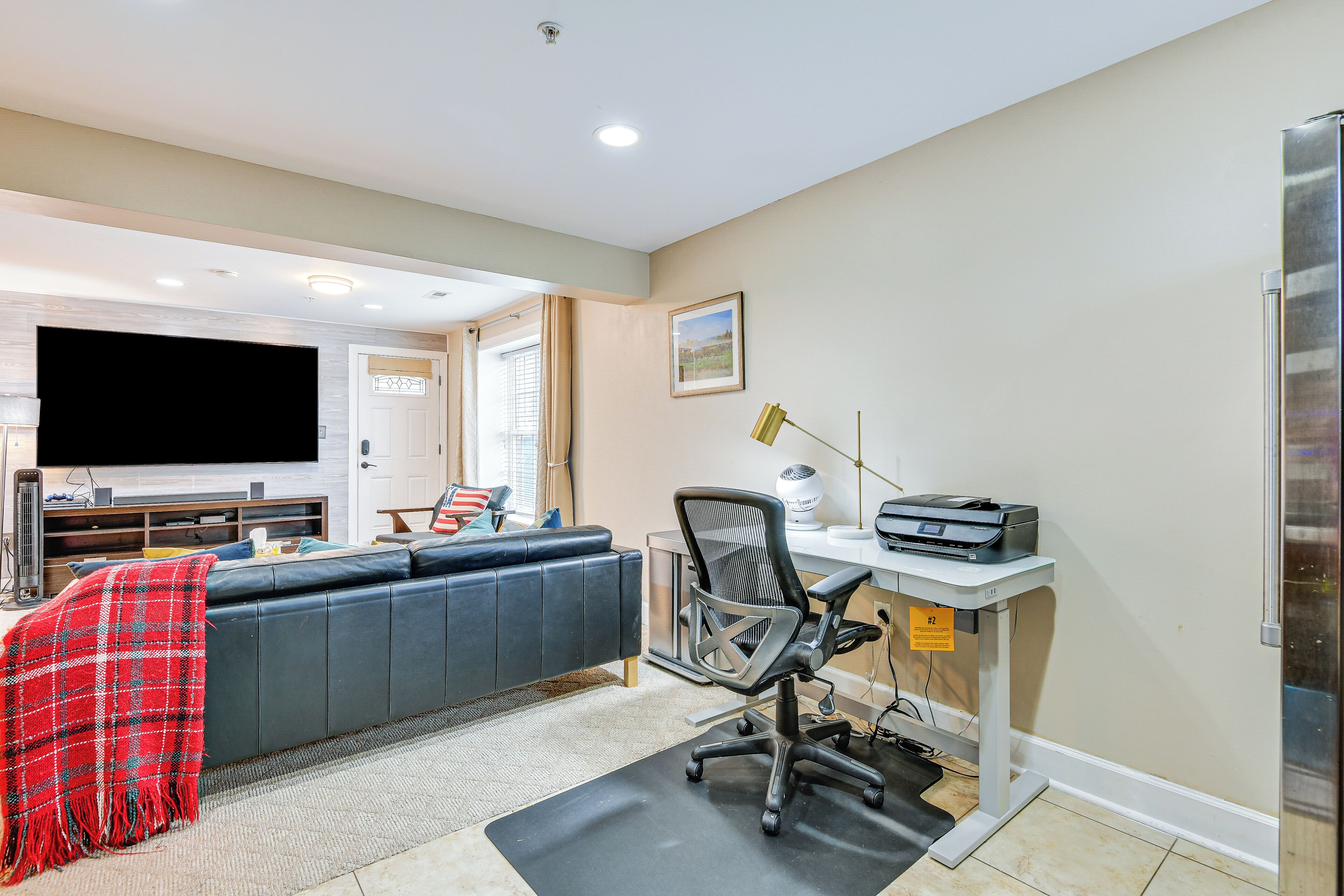 Een moderne woonkamer met een grote flatscreen televisie, een grijze bank, een bureau met een computer en een rode geruite plaid.