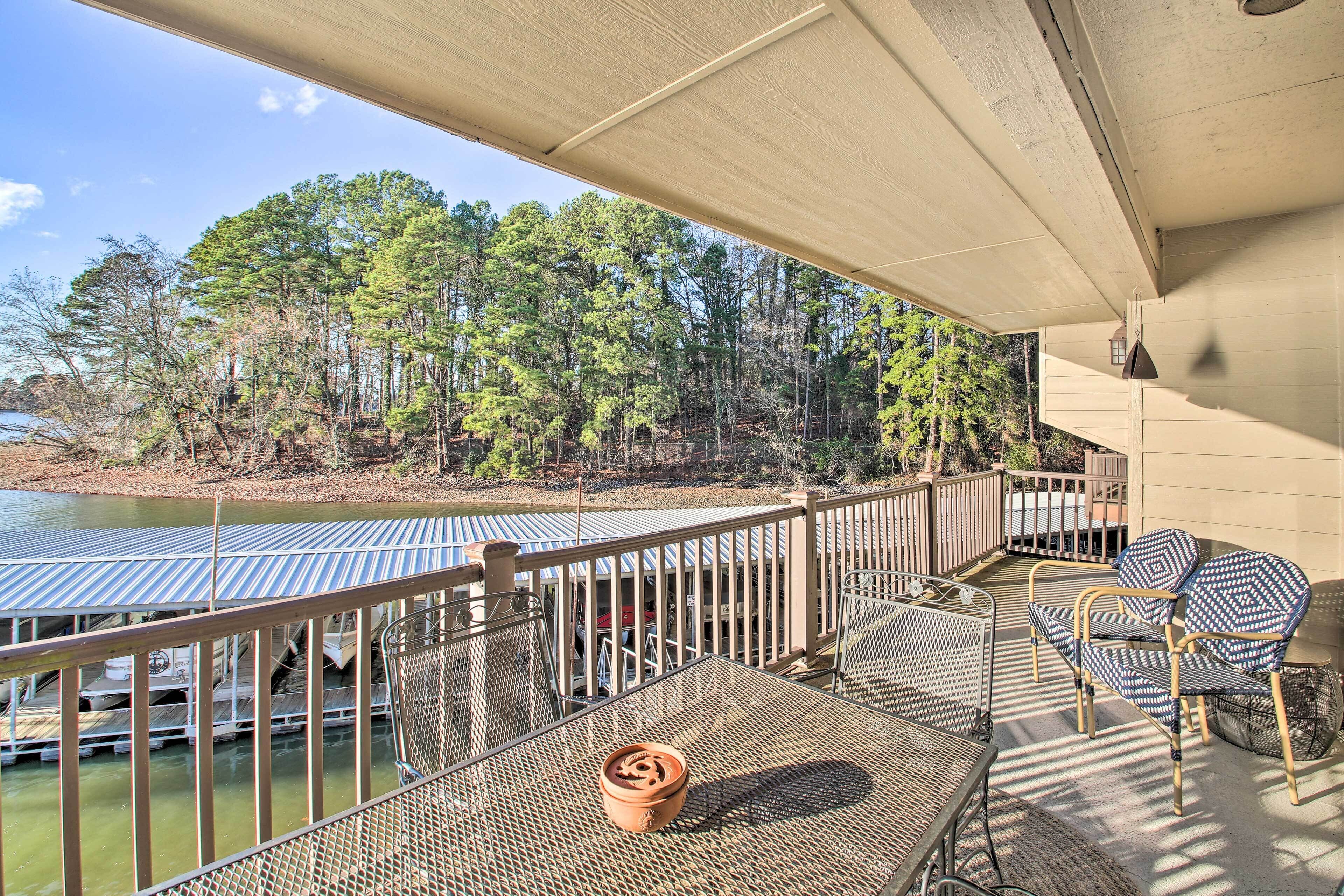 A covered outdoor area with a table and chairs, a metal railing, and a view of a forest and water body.