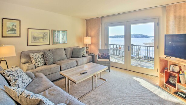 A living room with a sofa, coffee table, and a TV on a stand. There are framed pictures on the wall and a balcony with a view of the water. - Fireplace, Pool Access: Waterfront Hot Springs Gem (Hot Springs)
